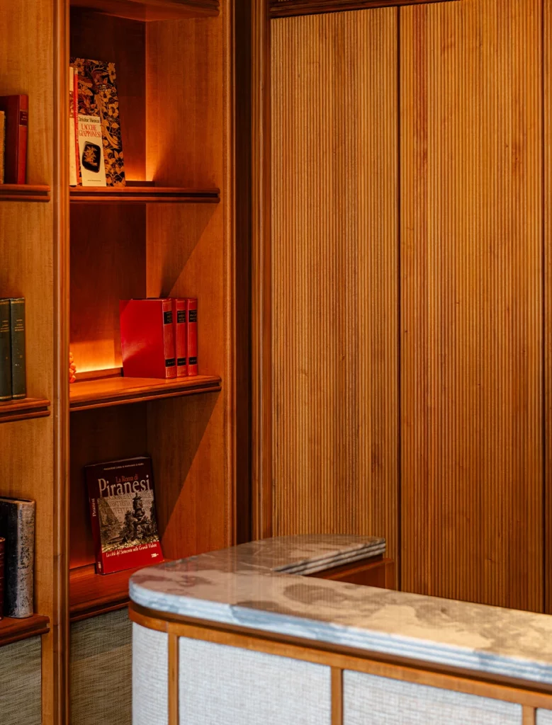Scaffalatura in legno scanalato con libri d’arte e bancone reception con piano in marmo.