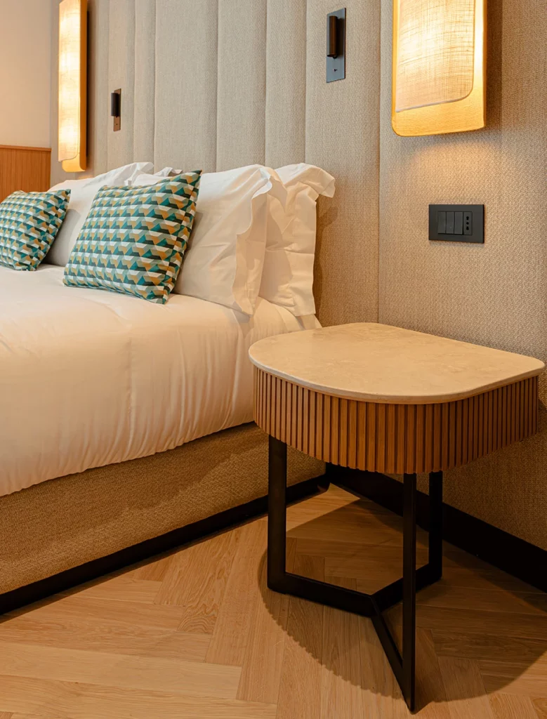 Comodino in legno con frontale scanalato accanto a letto matrimoniale con testiera imbottita e cuscini decorativi