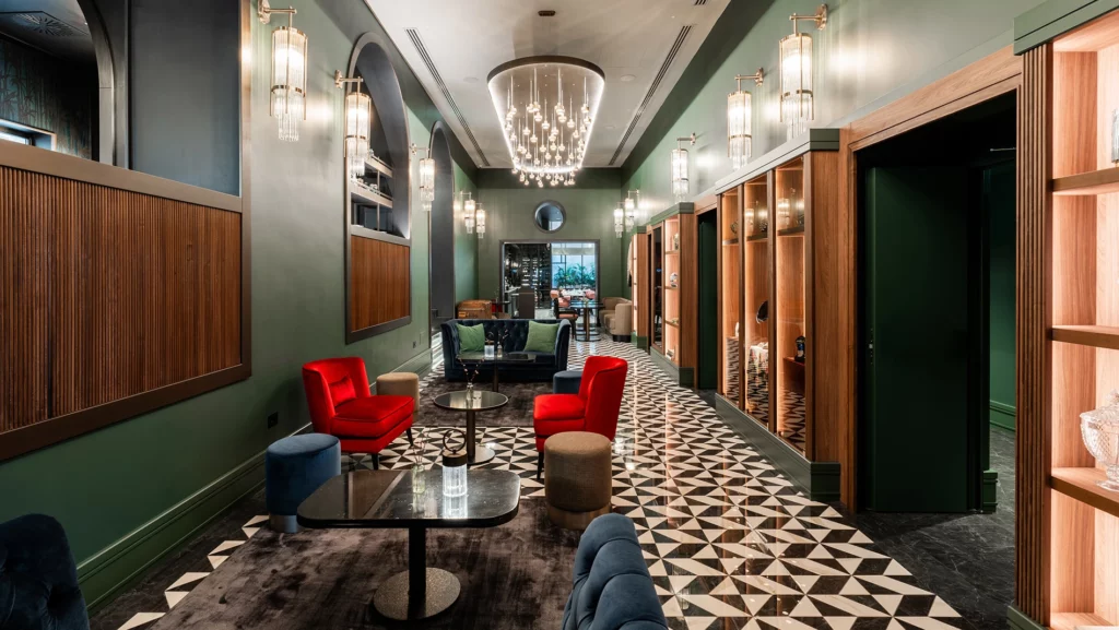 Lobby elegante con pareti verde scuro in legno, poltrone rosse e blu, lampadari in vetro e pavimento geometrico