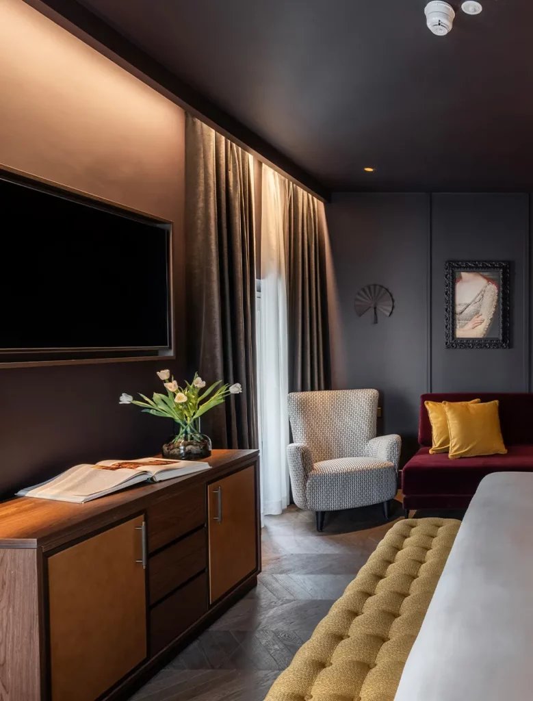 Camera d’hotel con arredi su misura, poltrona grigia e dettagli tessili color senape e bordeaux