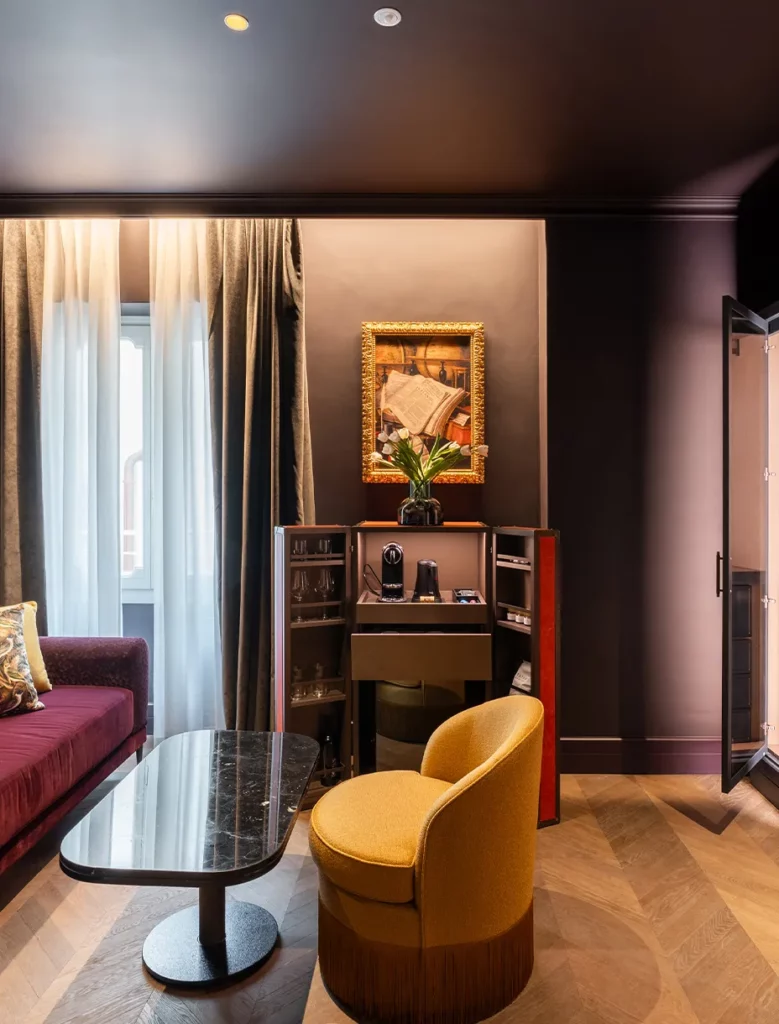 Zona living di suite con divano in velluto, poltrona gialla e minibar integrato con finiture di pregio.