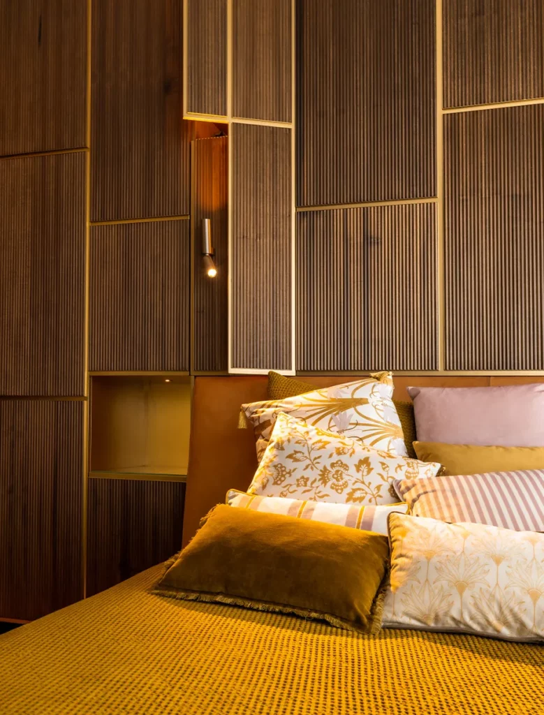 Dettaglio del letto con lenzuola dorate e cuscini. Armadio a scomparsa dietro la testiera de letto in legno