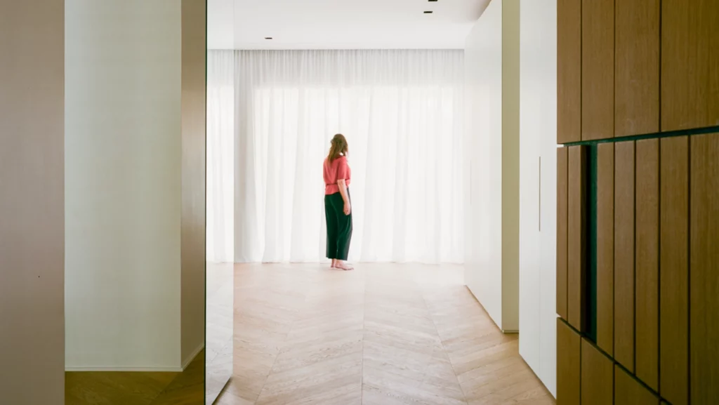 Ambiente residenziale con armadiature in legno su misura realizzata da Antonacci falegnamerie, pavimento in legno a spina di pesce e luce naturale filtrata da tende bianche con persona in piedi