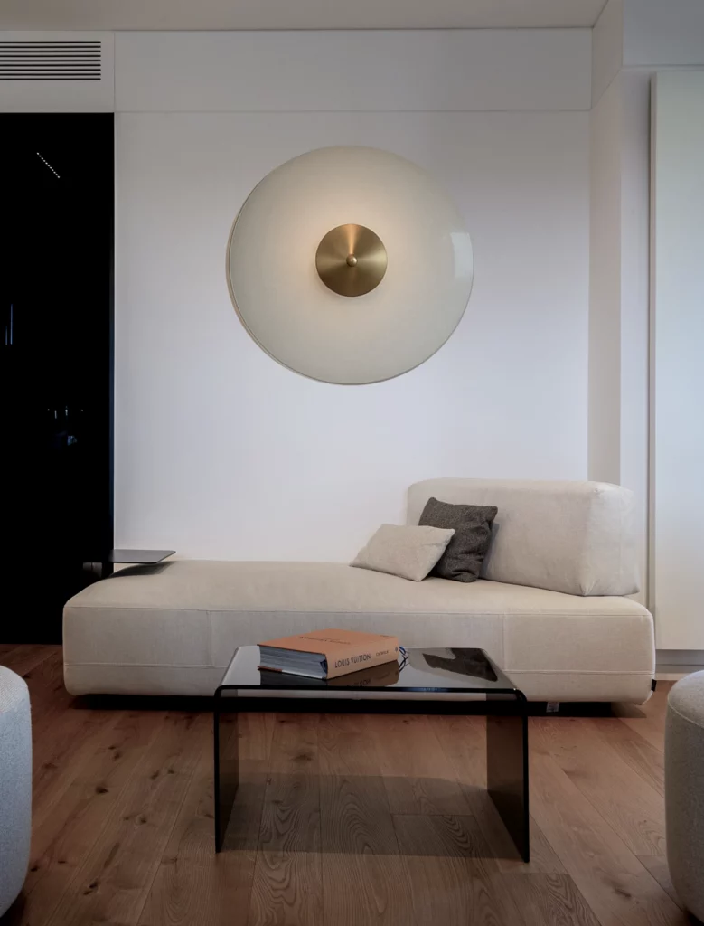 Soggiorno con divano moderno color crema, tavolino in vetro e applique circolare a parete su parquet Antonacci falegnamerie